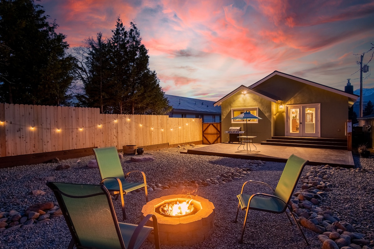 An outdoor seating area is featured, with four chairs arranged around a circular fire pit. The background shows a home with a deck area and a barbecue grill, under a painted sky at dusk. String lights are visible along the fence.