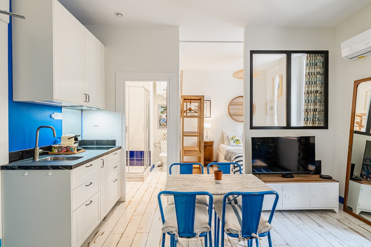 A compact kitchen area features sleek white cabinetry paired with a dark countertop. A dining table is set with four blue chairs, creating a cheerful dining space. A Smart TV is positioned against the wall, while a doorway leads to a separate living area and bathroom.
