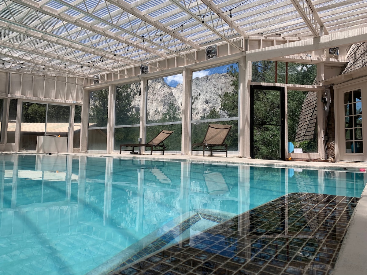 An indoor hot springs mineral pool is visible, surrounded by glass walls that offer views of mountains and trees. Two lounge chairs are positioned nearby on a deck, creating a serene atmosphere for relaxation.