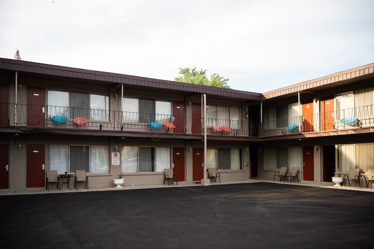 An exterior view of a two-story motel showcases multiple rooms with individual entrance doors leading to small balconies. Colorful chairs adorn the balconies, and a paved area is visible in the foreground. Nearby greenery adds a touch of nature.