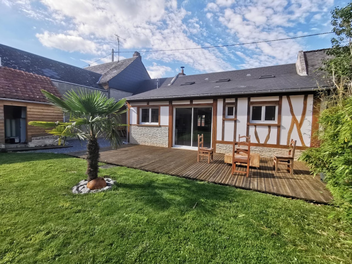 A terrace is visible, featuring wooden decking and a small palm tree. A seating area is arranged with wooden chairs. The exterior displays a mix of traditional and modern architectural elements, complemented by a grassy lawn and pebbled accents.