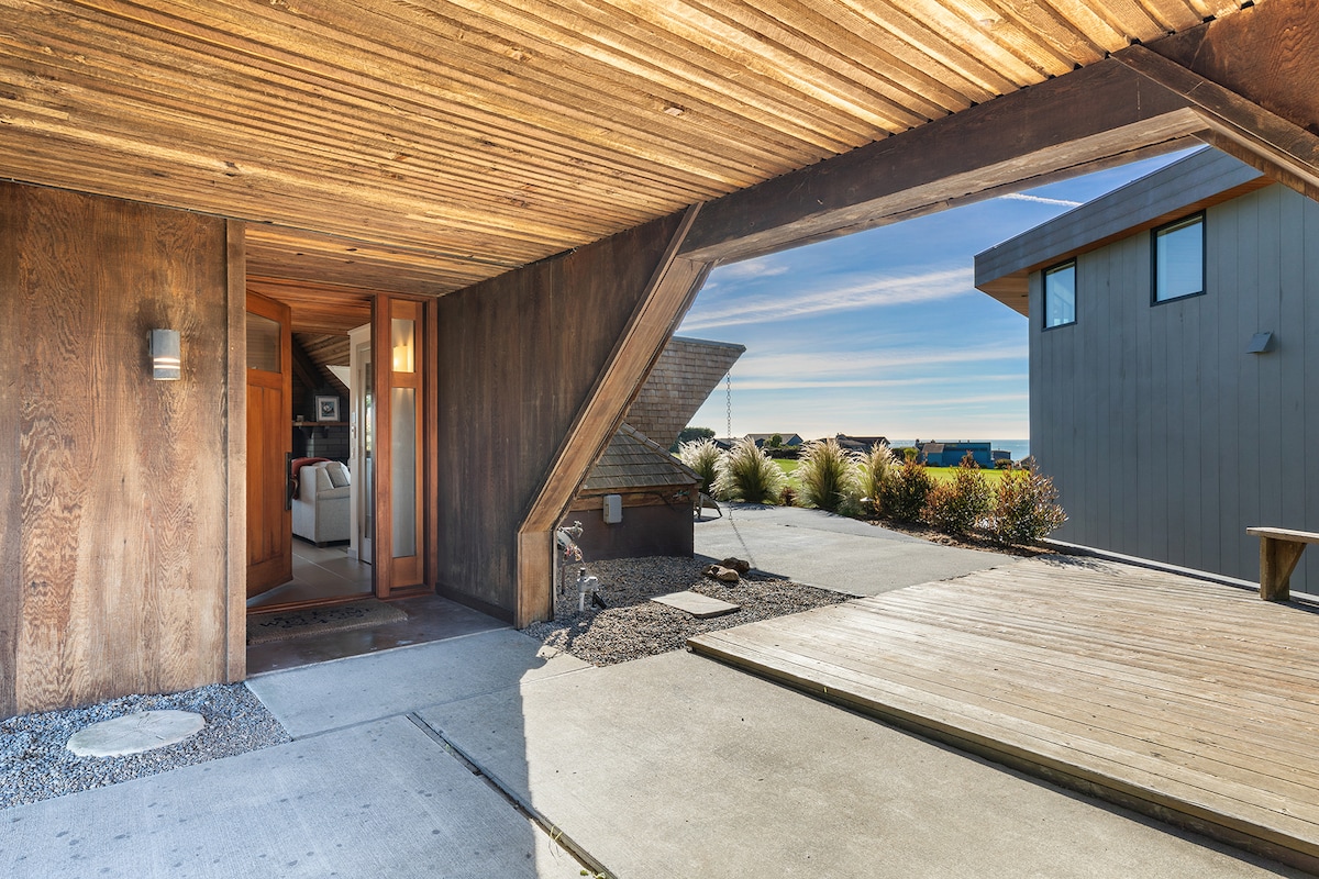 The entrance to the modern home is framed by unique architectural elements, featuring wooden paneling and clean lines. Natural light illuminates the entryway, offering glimpses of coastal views beyond. The outdoor space includes a paved pathway and a wooden deck area, enhancing the welcoming atmosphere.