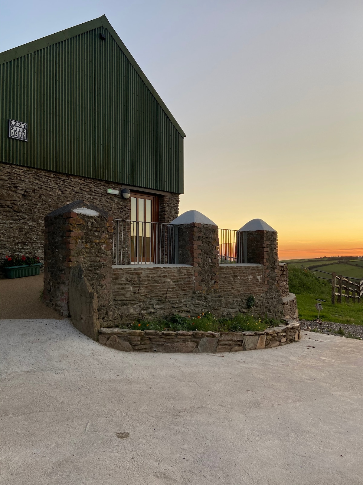 The exterior of the barn conversion is shown, featuring a rustic stone wall and a landscaped pathway leading to the entrance. The green metal roof contrasts with the warm tones of the sunset in the background, enhancing the structure's inviting presence.