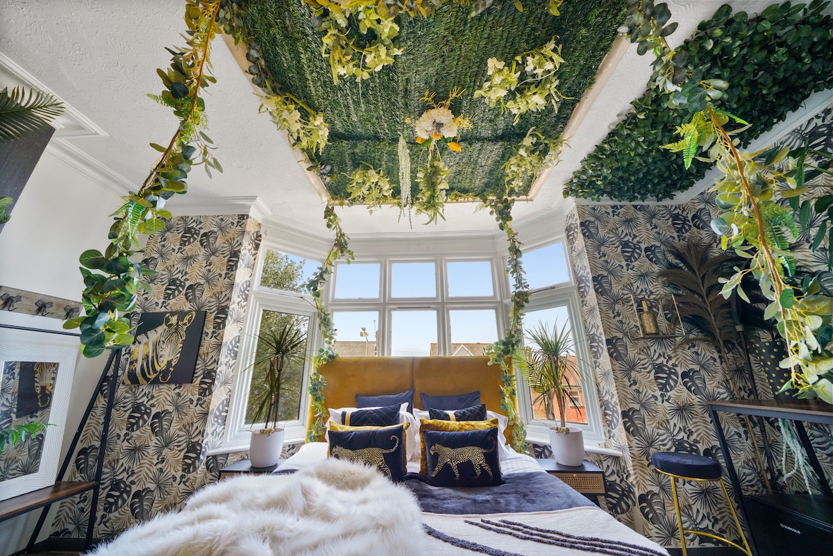 A vibrant bedroom is surrounded by botanical decor, featuring a ceiling adorned with lush greenery. Large windows allow natural light to fill the space, while a plush bed is complemented by decorative pillows and a cozy throw. Stylish wallpaper adds a unique flair to the room.
