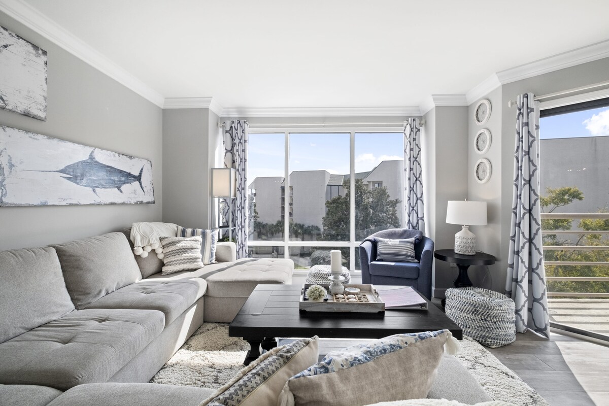 The living area features a spacious sectional couch, complemented by a dark wooden coffee table and a cozy rug. Floor-to-ceiling windows allow natural light to fill the room, offering views of the surrounding landscape. A blue accent chair adds a pop of color to the neutral decor.