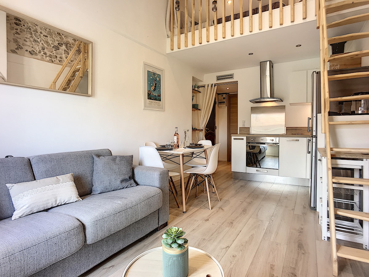 A modern main living area features a gray sofa with decorative pillows, a dining table for six, and a well-equipped kitchen. Natural light fills the space, highlighting the wooden staircase leading to the mezzanine. The decor blends rustic elements with contemporary design.