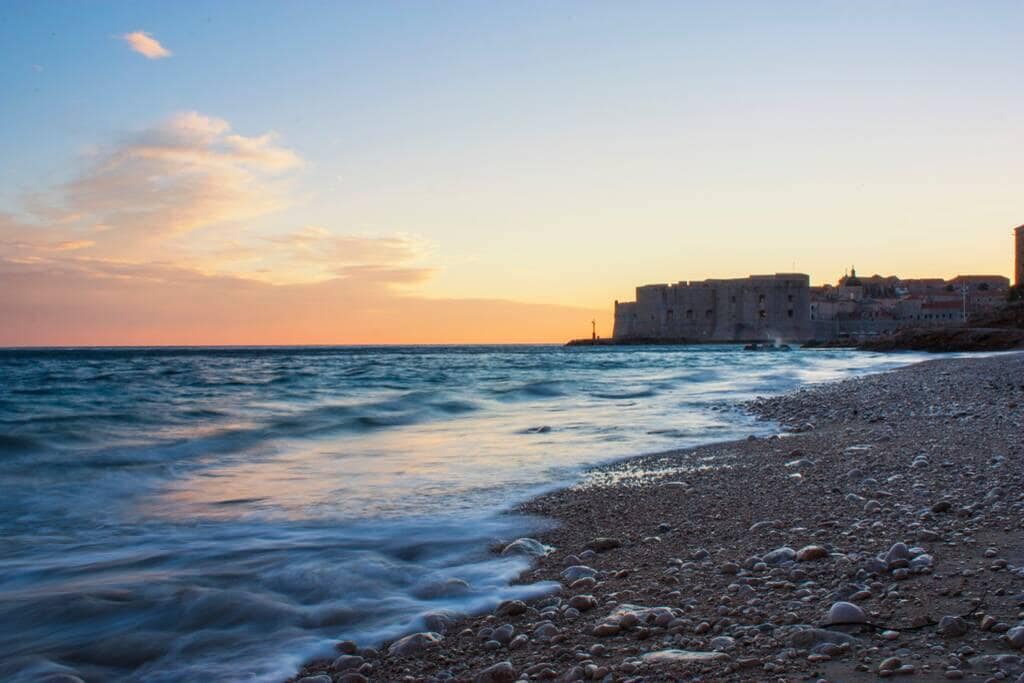 A serene beach scene captures gentle waves lapping at a shoreline adorned with smooth pebbles. In the distance, historic stone buildings are silhouetted against a colorful sky at sunset, transitioning from warm hues to cooler shades as day turns to night.