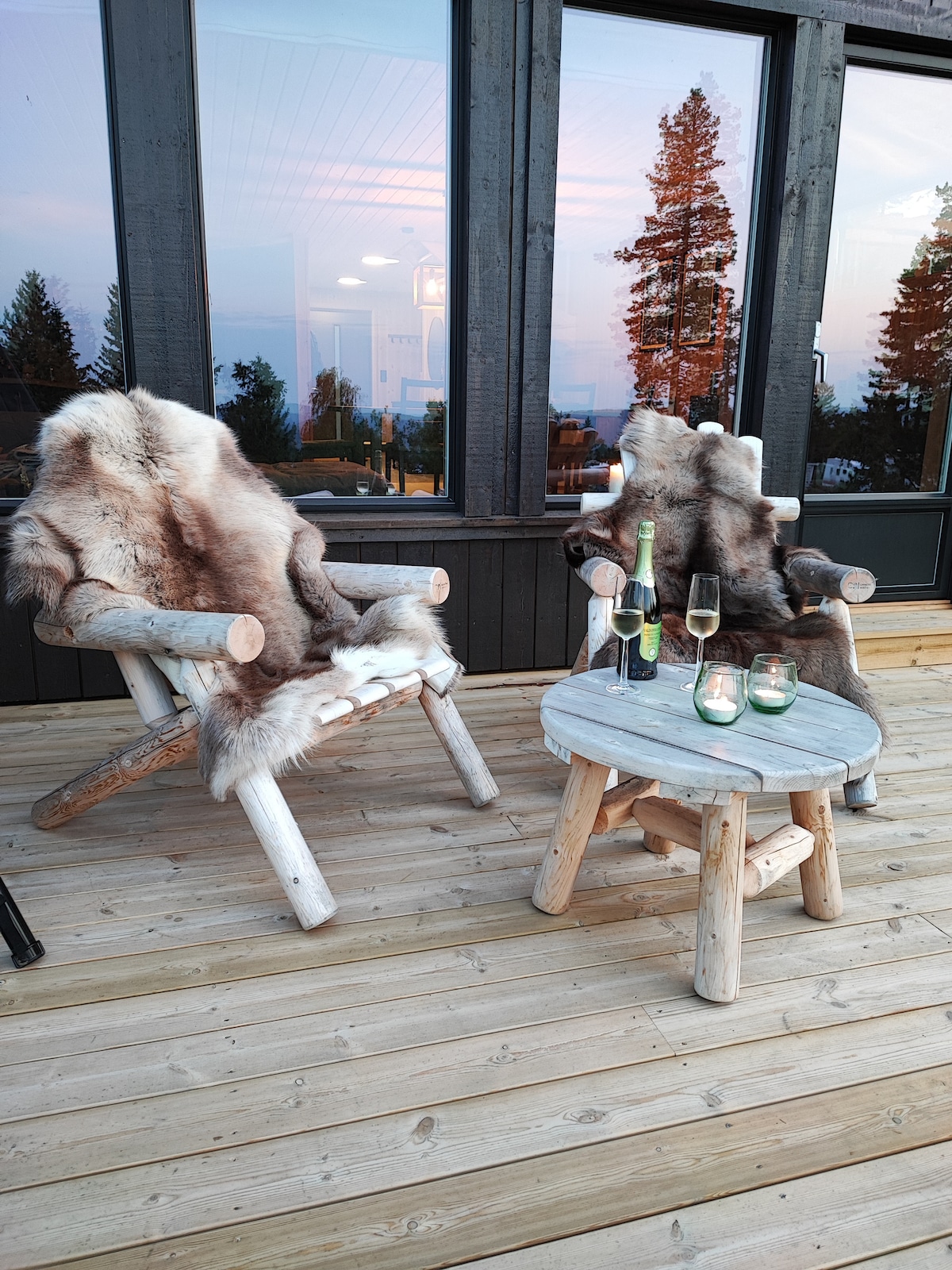 Two rustic chairs adorned with fur throws are positioned on a wooden deck, accompanied by a round table displaying a bottle of champagne and glasses. The scene is framed by large windows, with a backdrop of trees and a soft, colorful dusk sky.