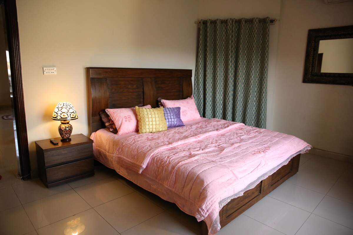 A spacious bedroom features a large bed with a soft pink quilt and decorative pillows. A bedside lamp adds a warm touch, while patterned curtains frame the window. The tiled floor reflects natural light from the room's entrance, enhancing the overall layout.