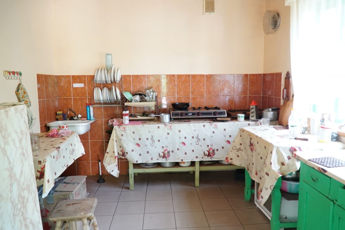 A shared kitchen is equipped with essential appliances and utensils. Counters are covered with patterned tablecloths, and dishware is stored on shelves. The space is illuminated by natural light from nearby windows, creating a functional area for meal preparation.