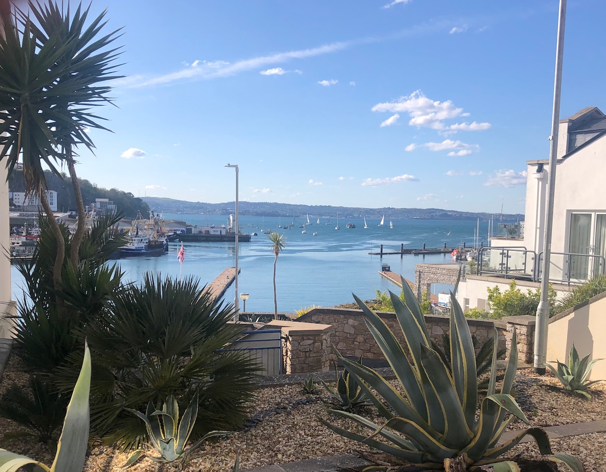 A view of the harbor is presented, showcasing calm waters dotted with sailboats. Lush plants frame the foreground, while buildings line the waterfront. In the distance, rolling hills create a serene backdrop under a clear blue sky with scattered clouds.