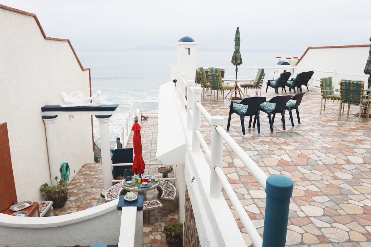 The outdoor terrace presents a spacious area with stone flooring, furnished with various seating arrangements. Tables and chairs are positioned strategically, overlooking the ocean. Colorful umbrellas provide shade, and vibrant potted plants add greenery to the setting.