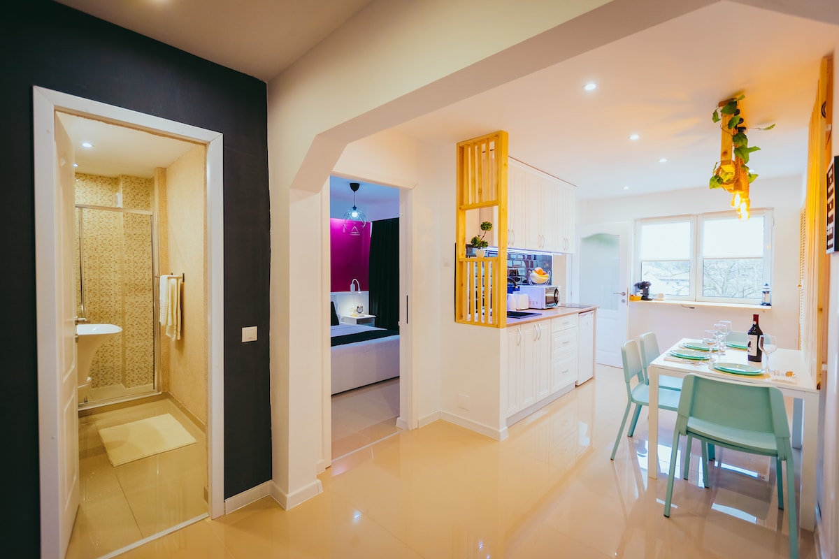 A modern kitchen area is visible, featuring a dining table with light green chairs set for a meal. A hallway leads to a bathroom with a shower, and views through the adjacent windows reveal natural light illuminating the space.