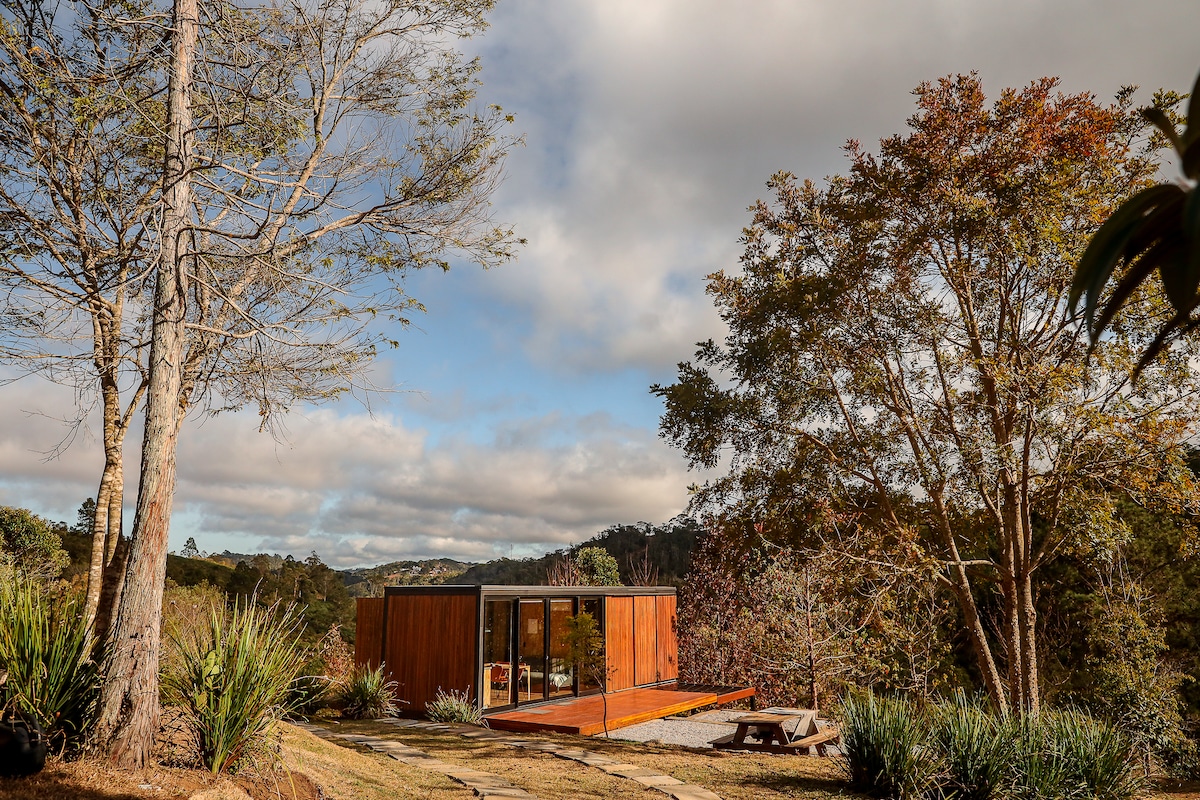 A minimalist cabana is set against a backdrop of lush greenery and cloudy skies. Floor-to-ceiling windows reflect the natural surroundings, while a picnic table nearby provides an outdoor gathering space. The landscape is punctuated by trees that enhance the serene atmosphere.