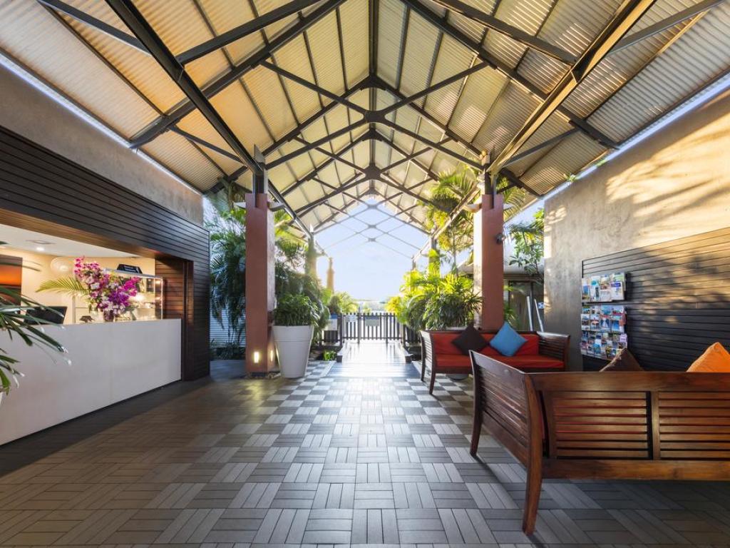An airy reception area is showcased, featuring a high, vaulted ceiling supported by beams. Comfortable wooden seating is arranged along the sides, with cushions adding a touch of color. Lush greenery is present, accompanied by a view of the outdoors through the open entrance.