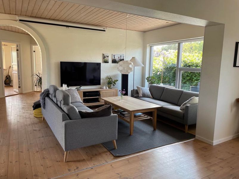 A spacious living area features two modern gray sofas arranged around a central wooden coffee table. A flat-screen TV is mounted on the wall, with plant decor providing a natural touch. Large windows allow natural light to flood in, framing views of the outdoor greenery.