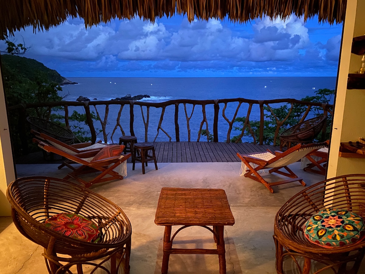 A cozy outdoor seating area showcases woven chairs and a wooden table, set against a backdrop of the ocean. The space is sheltered by a thatched roof, with tranquil views of the water and clouds reflecting the evening light.