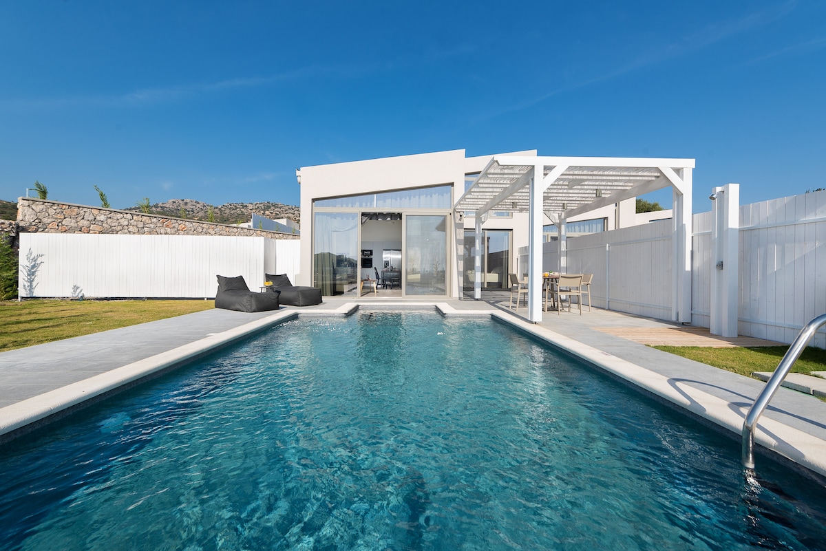 An outdoor area features a clear, inviting pool, framed by a simple patio. Behind the pool, large glass doors lead to a bright living space, creating a connection between indoor and outdoor environments. Comfortable seating is positioned nearby, along with a dining set under a shaded pergola.