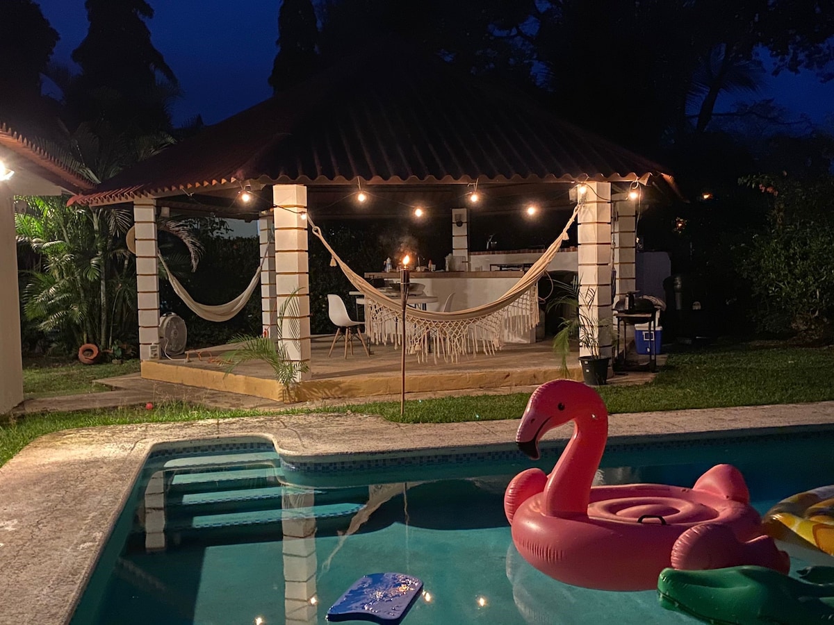 A charming outdoor space is highlighted by a covered gazebo featuring a dining area, surrounded by lush greenery. Soft lighting creates a relaxed ambiance. A swimming pool in the foreground includes colorful floaties, including a pink flamingo, enhancing the inviting atmosphere.
