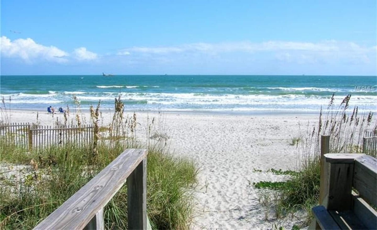 A sandy beach is visible, stretching towards the ocean under a clear blue sky. Gentle waves can be seen rolling onto the shore, with distant figures enjoying the sun. Tall grass and wooden railings frame the pathway leading to the beachfront.