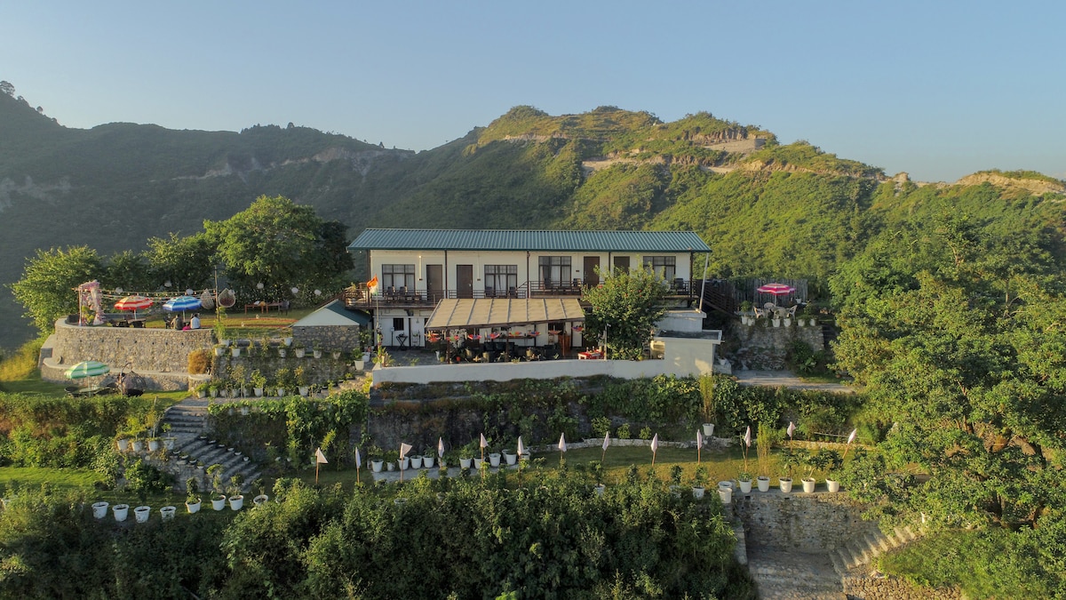 The hillside property features a two-story building surrounded by lush greenery and a landscaped garden. Outdoor seating areas with umbrellas are visible, alongside steps leading to the lawn adorned with white chairs, offering panoramic views of the surrounding mountains.