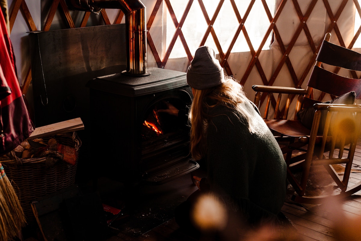 A cozy yurt interior is showcased, highlighting a wood stove with warm flames. A person is seated close to the stove, wearing a beanie. Surrounding elements include wooden rocking chairs and a basket, contributing to a comfortable and inviting atmosphere.