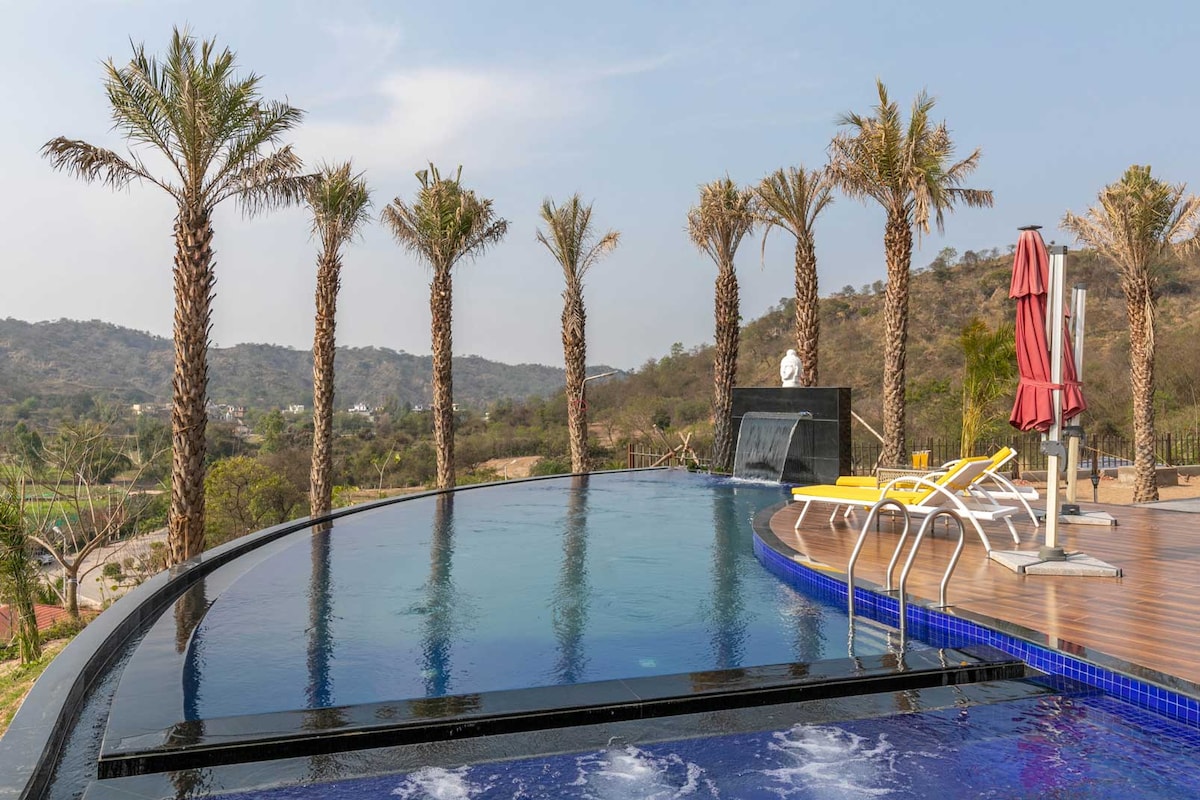 An infinity pool extends into the horizon, surrounded by palm trees. The sleek design features a seamless edge, while lounge chairs are placed nearby for relaxation. Gentle splashes can be seen, adding a serene ambiance to the outdoor space with hill views in the background.