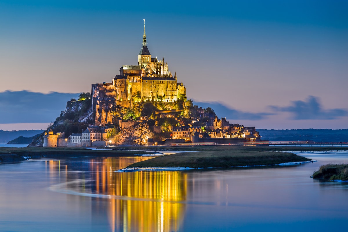 A historic island is depicted at dusk, with illuminated structures atop a rocky formation. The surrounding water reflects golden lights, enhancing the serene ambiance. The sky transitions from blue to a deeper hue, adding to the tranquil evening scene.