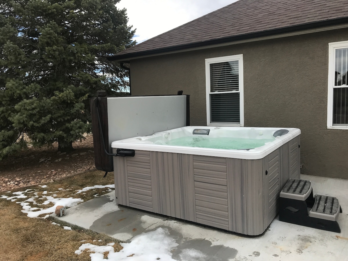 A large hot tub sits on a concrete patio surrounded by grass and scattered snow patches. It is situated against the side of a home, featuring steps for easy access. Windows from the house provide natural light to the area, and a tree is visible in the background.