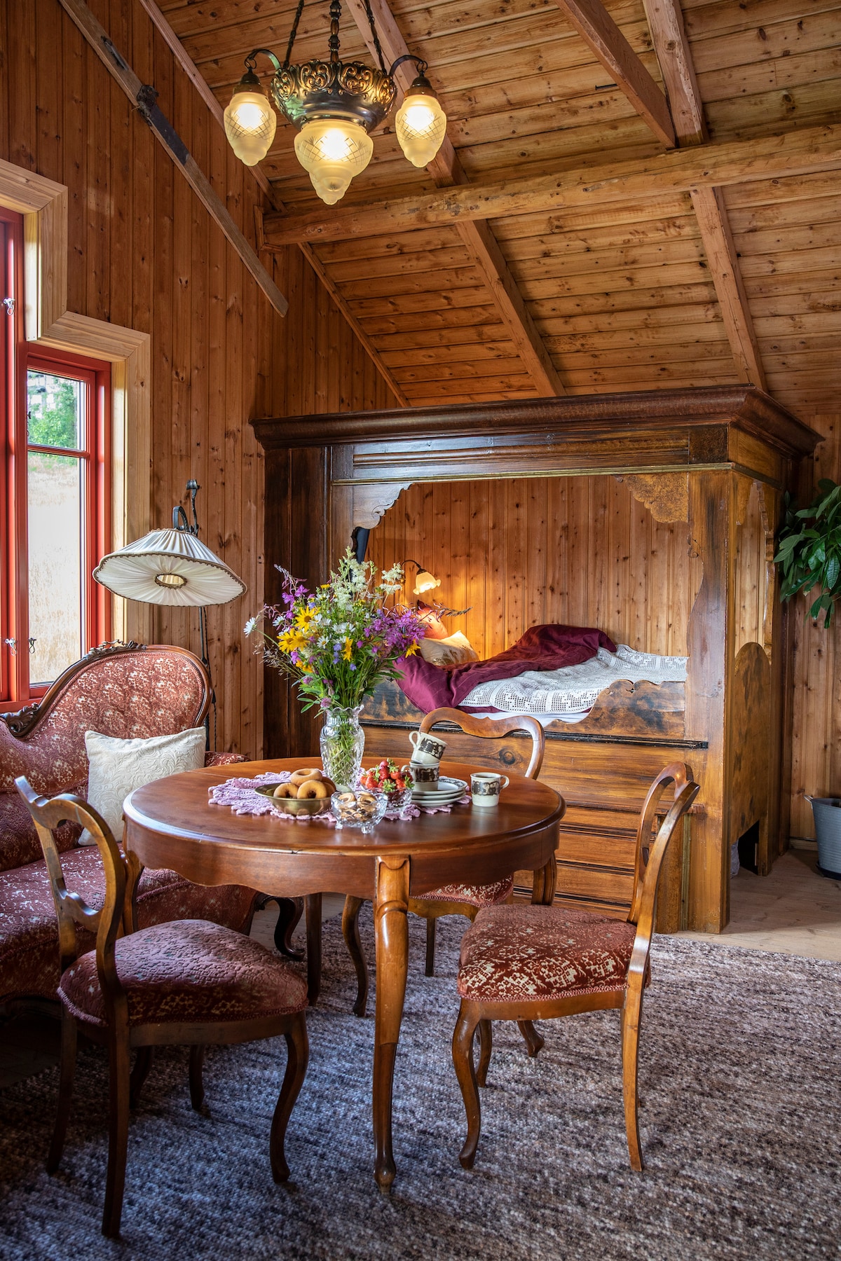 The interior showcases a wooden ceiling and warm wood paneling. A cozy dining area features a round table surrounded by four patterned chairs. A floral arrangement sits in the center, and a traditional bed with a canopy is situated against the wall, softly lit by ambient lighting.