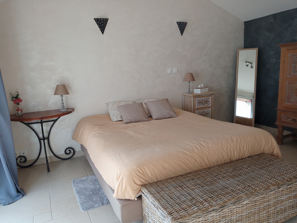 The bedroom features a spacious queen-size bed with soft bedding and two decorative pillows. A bedside table with lamps on either side is visible, along with a woven bench at the foot of the bed. A mirror reflects the room's neutral tones and textures.
