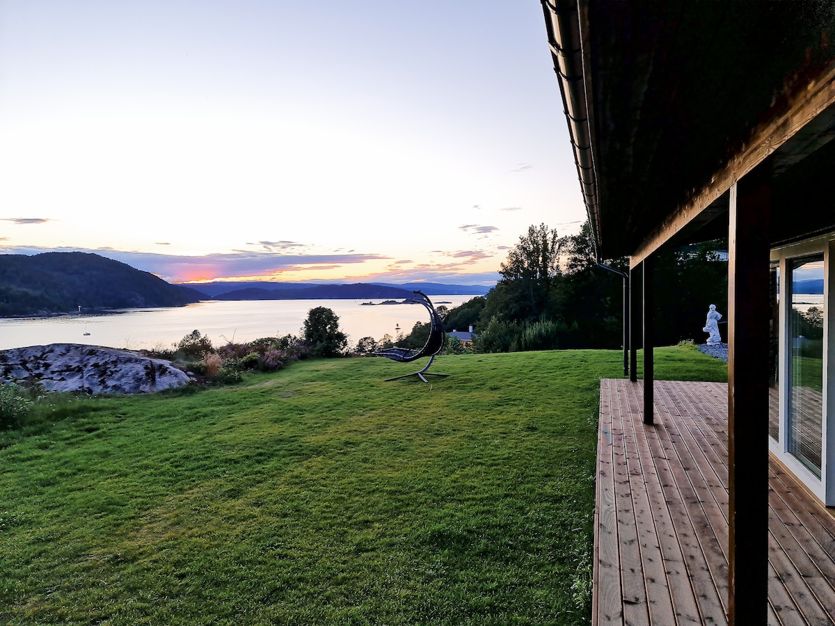 An outdoor view captures a spacious lawn lined with a wooden deck, leading toward a tranquil body of water. A sculptural piece is visible on the grass, framed by gently sloping hills and a colorful sky during sunset.