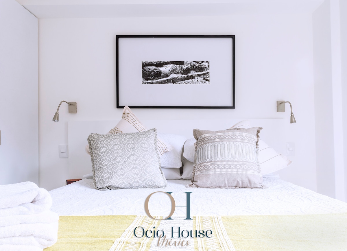 A cozy sleeping area features a spacious bed adorned with decorative pillows and a light textile spread. Wall sconces provide gentle illumination, and a framed artwork is displayed above the bed, enhancing the peaceful ambiance of the space.