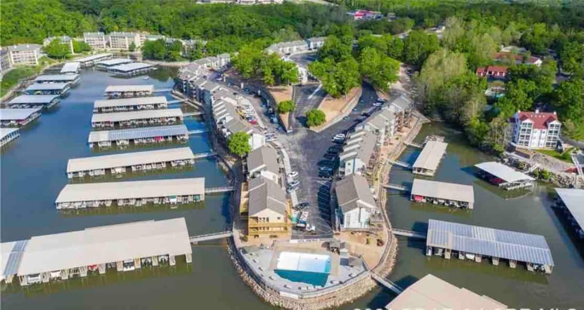 An aerial view captures the resort's layout, featuring multiple buildings surrounded by water. Boat docks with covered slips line the shoreline, and an inviting pool area is situated centrally, providing a glimpse of nearby green foliage and the expansive lake beyond.