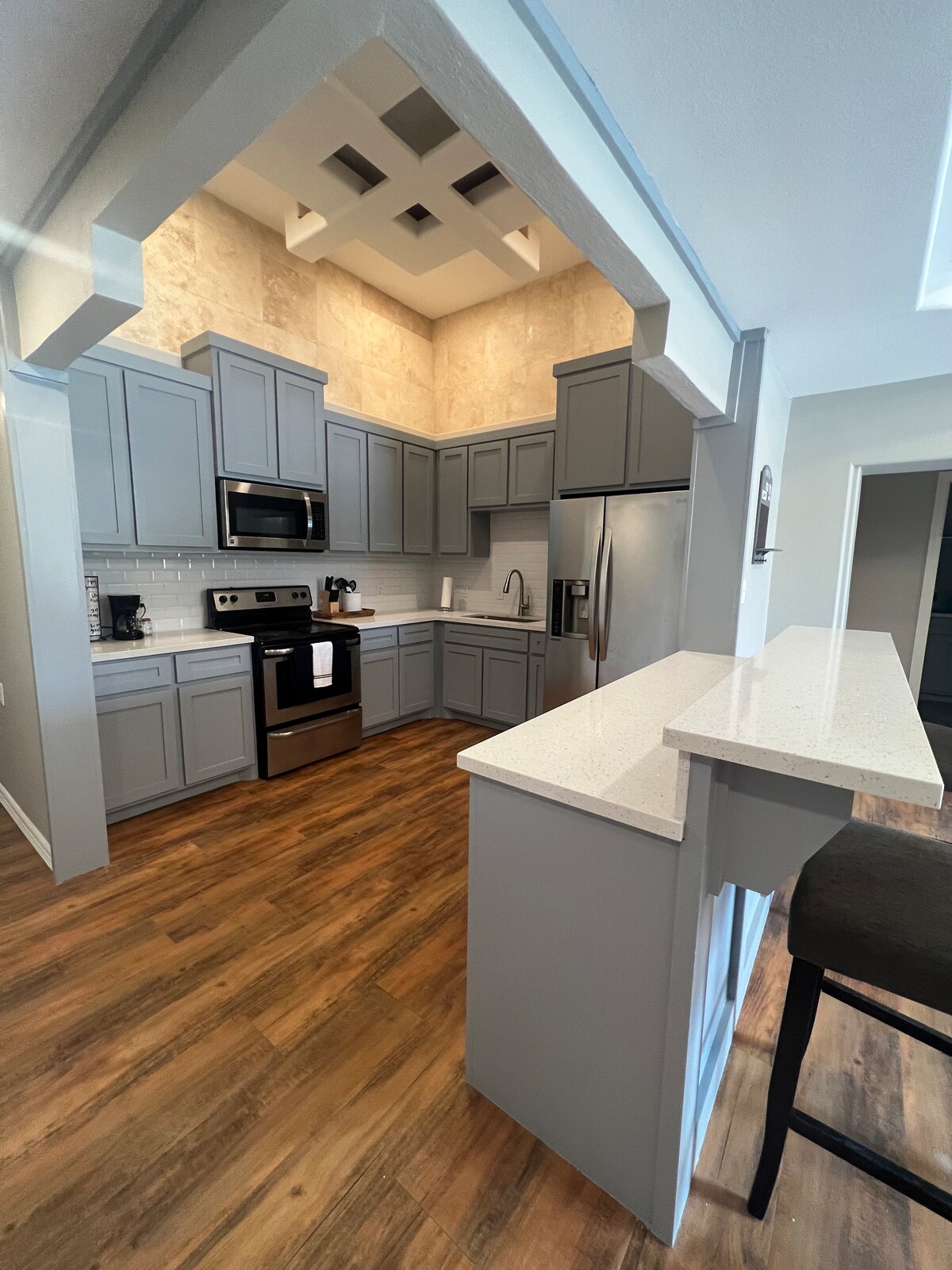 A newly renovated kitchen features modern gray cabinetry and stainless steel appliances. A bar with seating provides a convenient dining space. The open layout enhances natural light, highlighting the spacious counters and textured wall design.