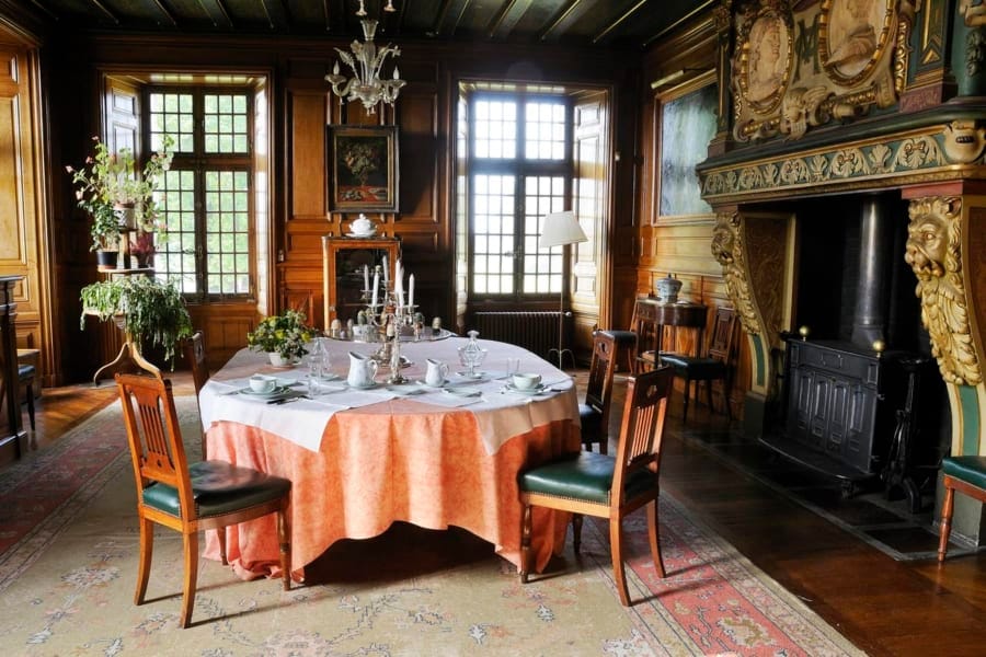 A family dining room is showcased with a large round table draped in a light-colored tablecloth, surrounded by wooden chairs. The elegant decor includes a chandelier and a fireplace, while large windows provide natural light, highlighting the rich wooden paneling and intricate detailing.