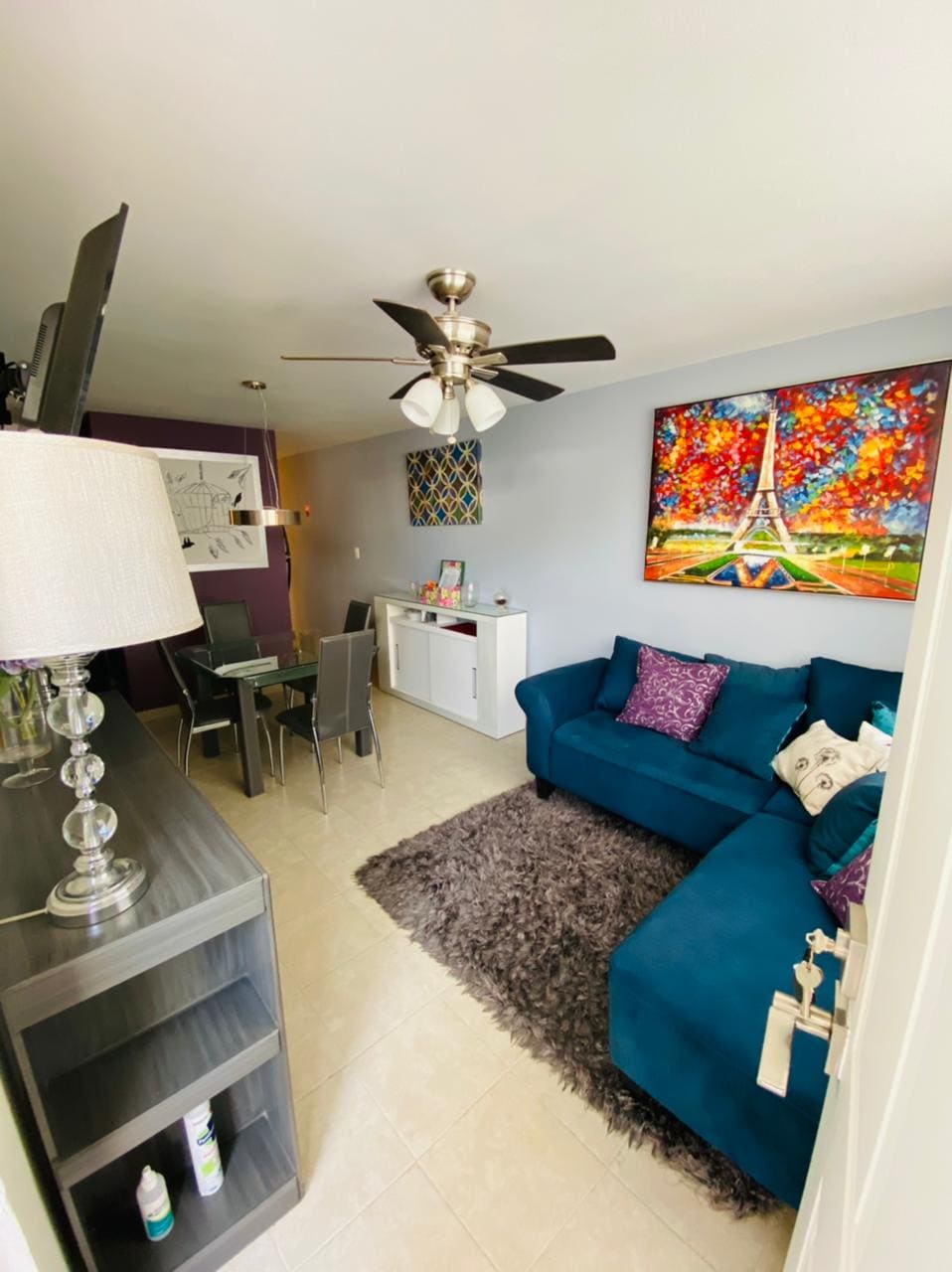 The living area features a blue sectional sofa set against a colorful artwork depicting the Eiffel Tower. A modern dining table with transparent chairs complements the space. A light rug adds texture, while a ceiling fan enhances comfort. Natural light illuminates the room.
