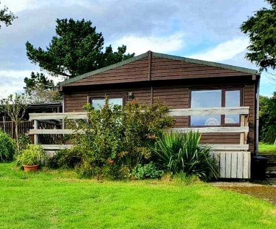 A wooden chalet exterior is framed by greenery, featuring a deck with a railing. Large windows provide views of the surrounding landscape. The structure is set amidst a grassy area that blends with the natural surroundings.