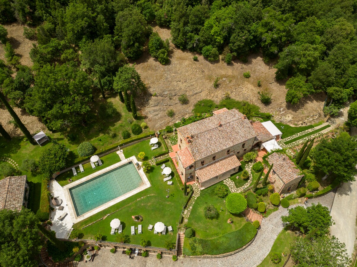 An aerial view of the property highlights a historic structure surrounded by lush greenery. A spacious swimming pool sits adjacent to the building, complemented by well-maintained gardens and patio furniture for relaxation. The natural landscape enhances the tranquil atmosphere of the retreat.