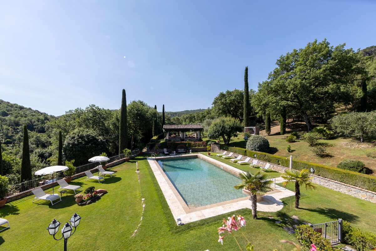 A serene outdoor pool area is surrounded by lush greenery, featuring several lounge chairs along the edge. Tall cypress trees and palm plants add to the tranquil landscape. A gazebo is visible in the background, providing a shaded area for relaxation.