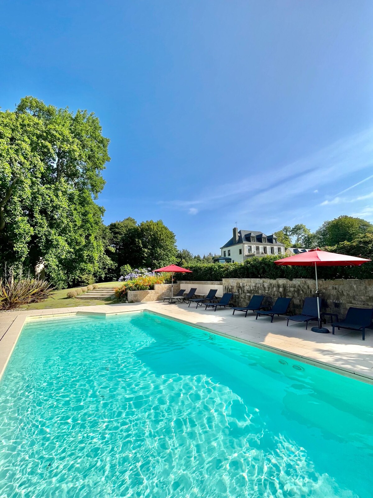 An outdoor swimming pool is surrounded by lounge chairs beneath bright red umbrellas. Lush greenery lines the pool area, and a white mansion is visible in the background, complementing the clear blue sky overhead.