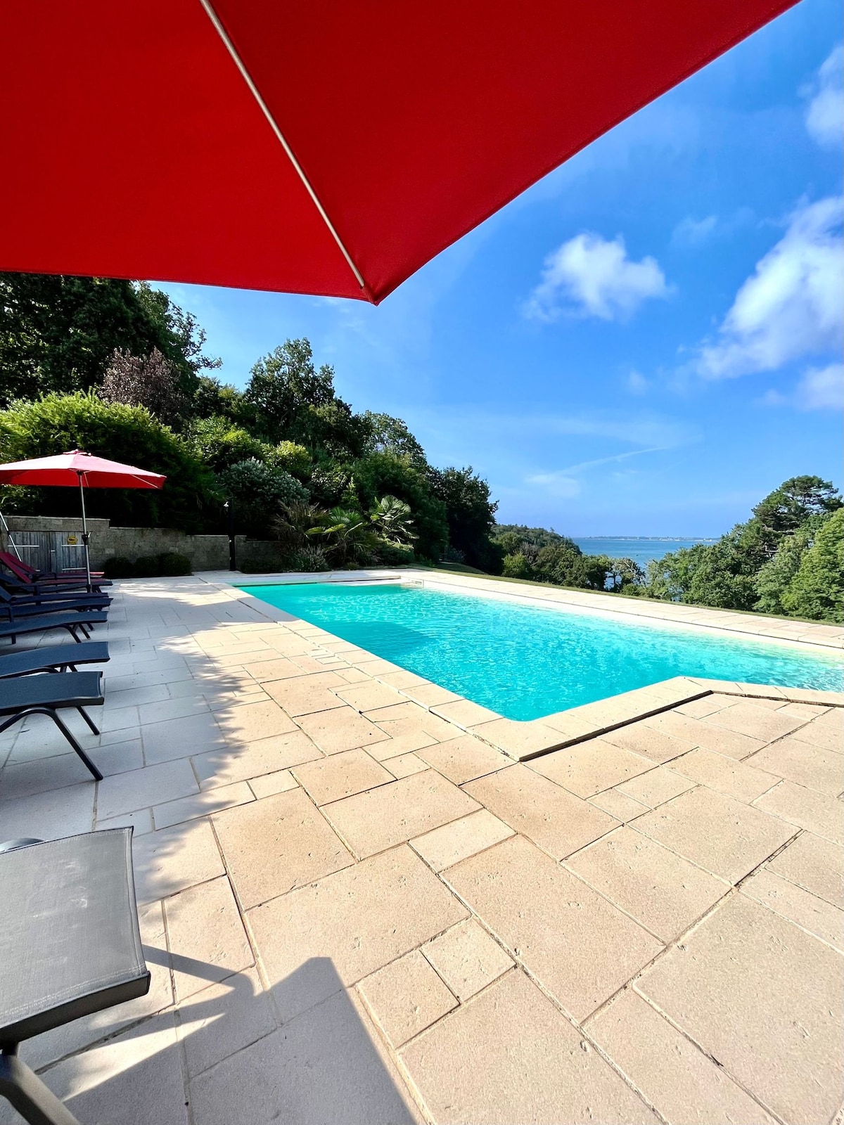 An inviting outdoor pool is surrounded by a spacious stone terrace, complete with several lounge chairs shaded by red umbrellas. Lush greenery frames the area, and a clear blue sky reflects off the serene water, offering a sense of tranquility and openness.