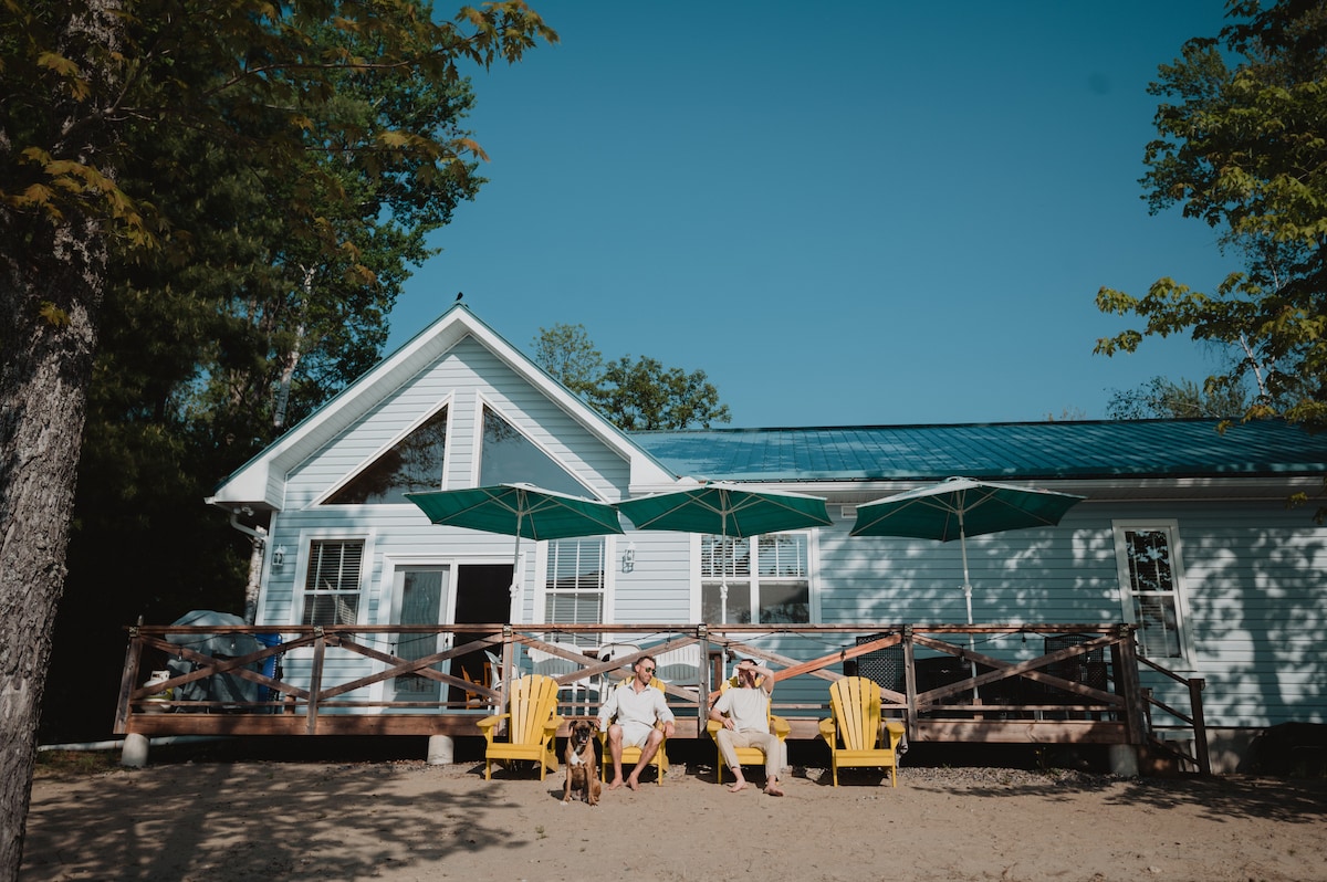 A light blue chalet features a spacious wooden deck with bright yellow chairs and two umbrellas. Two individuals are seated, accompanied by a dog. The sandy area and surrounding greenery create a welcoming outdoor setting under clear skies.
