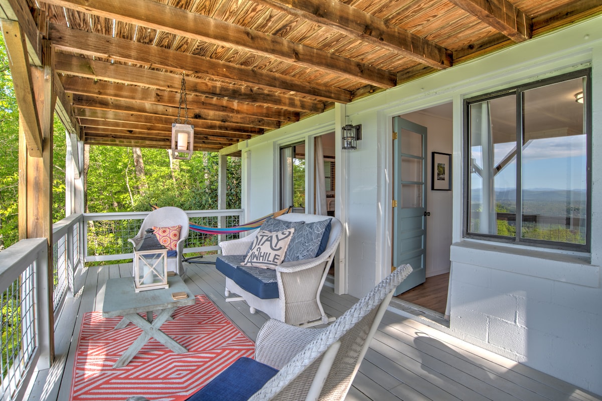 A covered deck is shown with a wooden ceiling and a blend of seating options, including a cozy wicker chair and a patterned lounge chair. A small table sits in the center, and large windows provide views of the surrounding greenery.