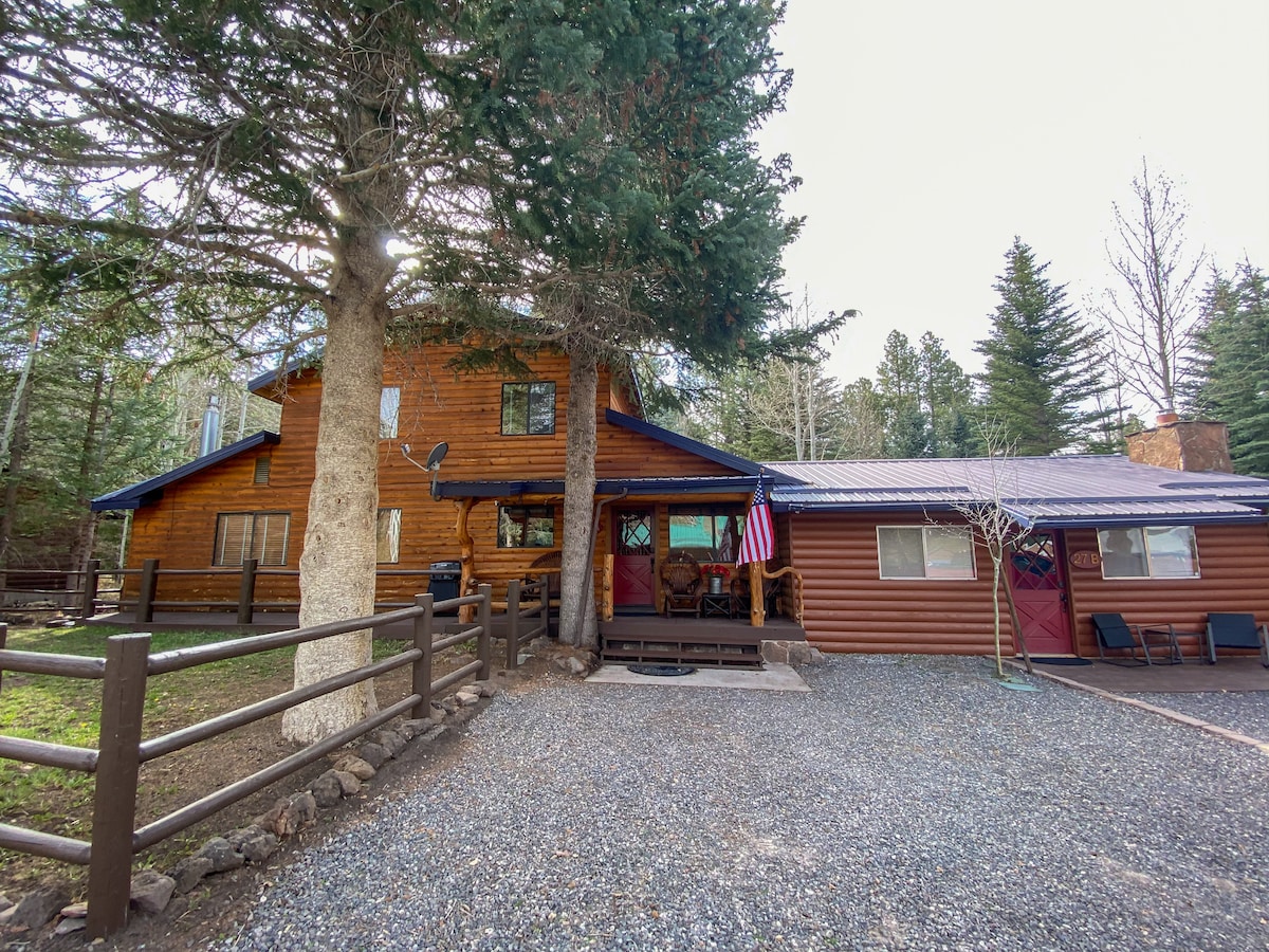 The exterior of a rustic cabin and cottage is shown, framed by tall trees. A gravel driveway leads to the entrance, where a porch with seating and an American flag are visible. The wooden structure features a combination of rich brown siding and a blue roof.