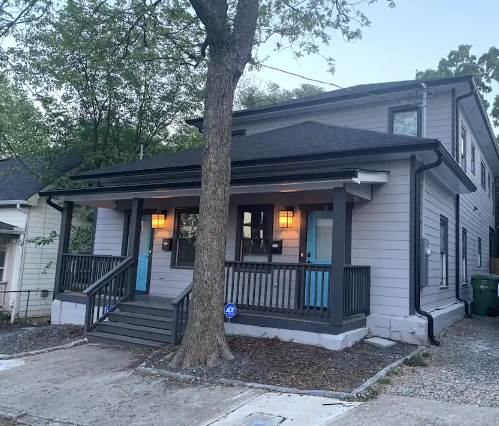 The exterior of the duplex features a welcoming entrance with two blue doors framed by stylish lanterns. A spacious front porch with railings is highlighted, while a large tree provides shade nearby. The surrounding area includes a gravel driveway and green foliage.