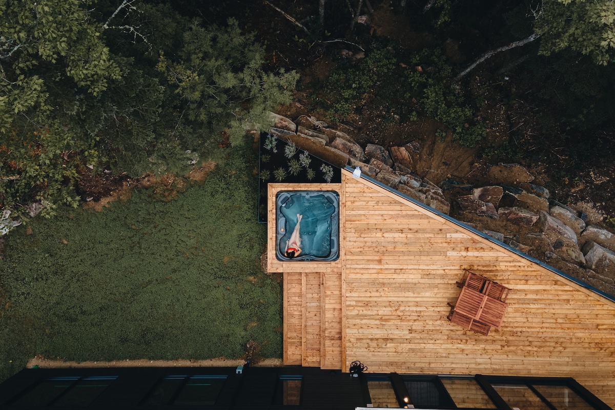 An aerial view showcases a wooden deck leading to a private hot tub, surrounded by lush greenery. A spacious seating area is available nearby, providing a serene outdoor space for relaxation amidst nature.