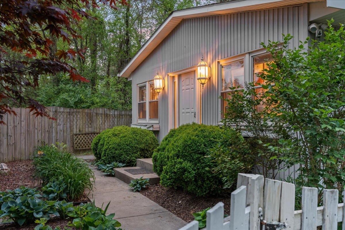 The exterior of a cozy retreat is displayed, featuring a well-maintained path leading to a welcoming entrance. Lush greenery and manicured hedges frame the structure, while outdoor lighting enhances the inviting atmosphere as dusk approaches.