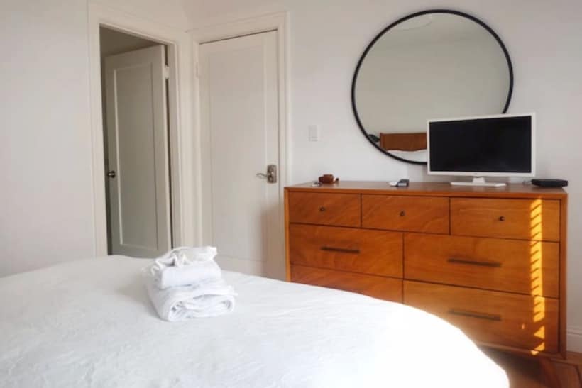 A spacious bedroom features a neatly made bed with fresh white linens, accompanied by stacked towels. A wooden dresser with multiple drawers is positioned beside a circular mirror. A smart TV is mounted on the wall, and a door leads to a private en-suite bathroom.