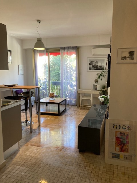 The living area features a small table with high chairs and a coffee table, surrounded by natural light from a large window with sheer curtains. Greenery outside is visible through the glass doors, and a decorative console table adds a contemporary touch.